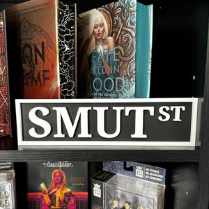 May include: A black shelf displays books, action figures, and a street sign. The books have colorful covers with titles like "On Game" and "A Fate Inked In Blood." Action figures of characters from "Buffy the Vampire Slayer" are visible, along with a "SMUT ST" sign.