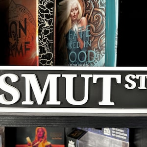 May include: A rectangular sign with the text "SMUT ST" in bold, white letters against a black background. The sign is framed in white and sits on a black shelf, with books visible in the background.