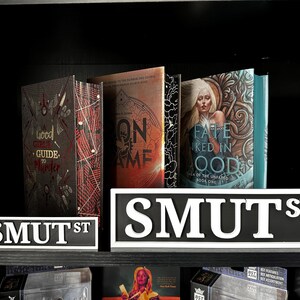 May include: A shelf displays several books with unique covers, including "Good Crime Guide to Murder" and "Fate Ked in Ood." Two "SMUT ST" signs are in the foreground. The books have various colors, including red, orange, and teal.