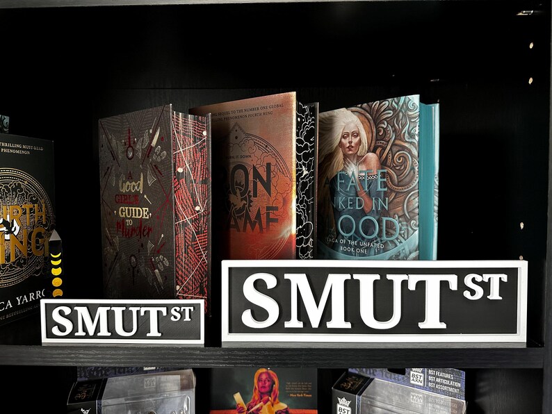 May include: A shelf displays several books with unique covers, including "Good Crime Guide to Murder" and "Fate Ked in Ood." Two "SMUT ST" signs are in the foreground. The books have various colors, including red, orange, and teal.