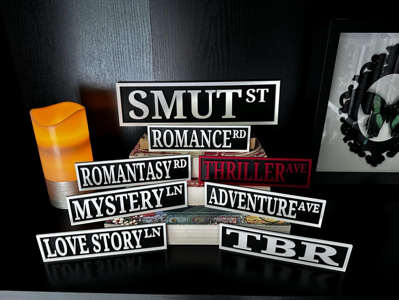 May include: A collection of black and white street sign-style plaques with various book genre names in bold, white lettering. The signs include "SMUT ST", "ROMANCE RD", "ROMANTASY RD", "MYSTERY LN", "LOVE STORY LN", "THRILLER AVE", "ADVENTURE AVE", and "TBR".