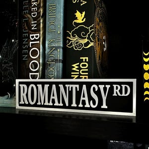 May include: A black and white sign that reads "ROMANTASY RD" in bold, white letters. The sign is on a shelf in front of a row of books with dark covers. The books have gold and white lettering.