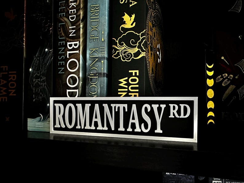 May include: A black and white sign that reads "ROMANTASY RD" in bold, white letters. The sign is on a shelf in front of a row of books with dark covers. The books have gold and white lettering.