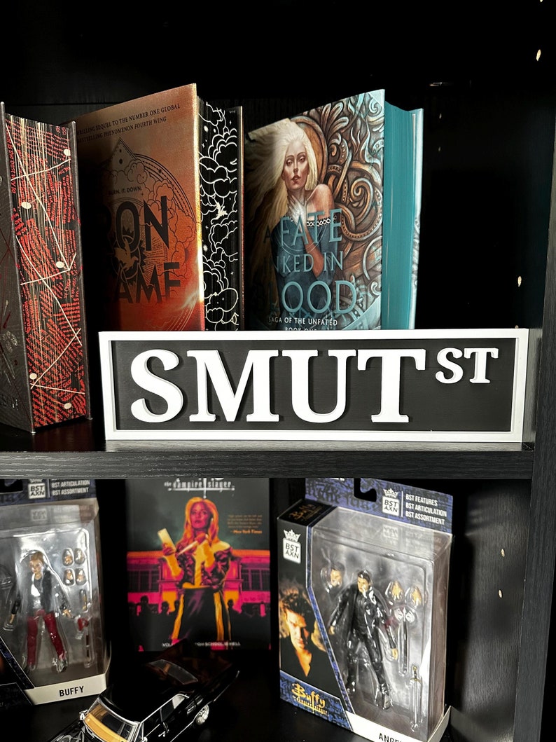 May include: A black shelf displays books, action figures, and a street sign. The books have colorful covers with titles like "On Game" and "A Fate Inked In Blood." Action figures of characters from "Buffy the Vampire Slayer" are visible, along with a "SMUT ST" sign.