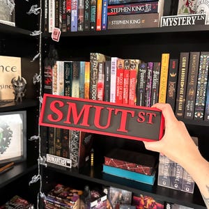 May include: A black and red "SMUT ST" sign is held in front of a bookshelf filled with books. The sign has red letters on a black background. Bookshelves are filled with various books, including Stephen King novels.