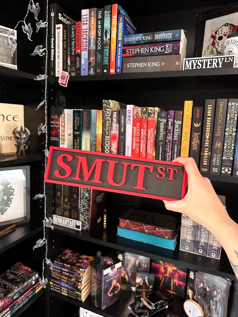 May include: A black and red "SMUT ST" sign is held in front of a bookshelf filled with books. The sign has red letters on a black background. Bookshelves are filled with various books, including Stephen King novels.
