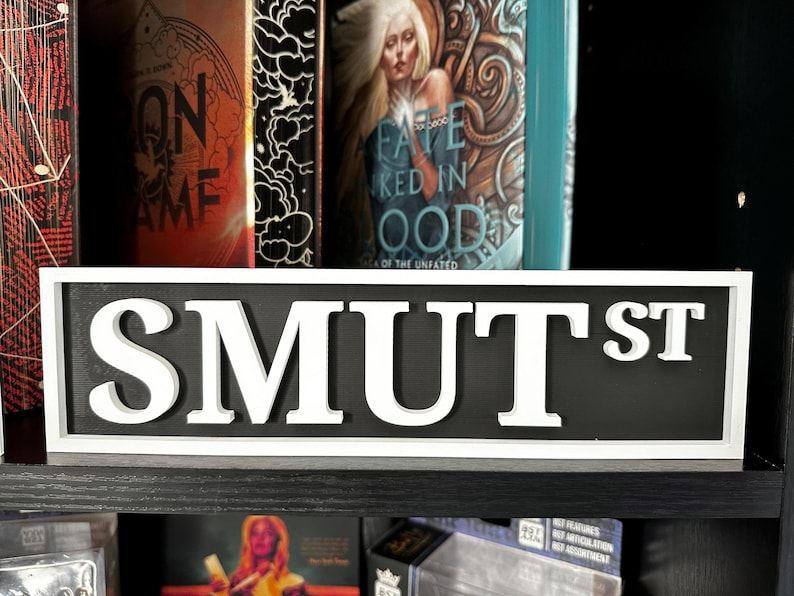 May include: A rectangular sign with the text "SMUT ST" in bold, white letters against a black background. The sign is framed in white and sits on a black shelf, with books visible in the background.