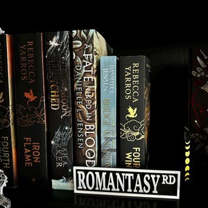 May include: A collection of fantasy books with titles like "Fourth Wing," "Iron Flame," and "A Fate Inked in Blood" on a black shelf. A street sign reading "Romantasy Rd" sits in front of the books. Fairy lights are draped on the left.