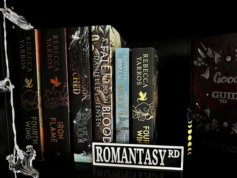 May include: A collection of fantasy books with titles like "Fourth Wing," "Iron Flame," and "A Fate Inked in Blood" on a black shelf. A street sign reading "Romantasy Rd" sits in front of the books. Fairy lights are draped on the left.