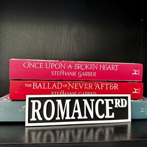 May include: A stack of books with red and teal covers. The top two books are titled "Once Upon a Broken Heart" and "The Ballad of Never After." A black and white sign that reads "ROMANCE RD" is in front of the books.