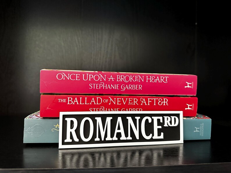 May include: A stack of books with red and teal covers. The top two books are titled "Once Upon a Broken Heart" and "The Ballad of Never After." A black and white sign that reads "ROMANCE RD" is in front of the books.