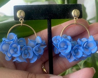 May include: A pair of gold-toned hoop earrings with clusters of light blue and gray floral accents. The earrings are displayed on a black stand, showcasing their design and color contrast. The floral elements add a delicate touch.