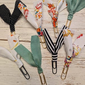 May include: A set of eight decorative paper clips with fabric embellishments. The clips are gold and black, and the fabric is a variety of patterns, including floral, polka dot, and striped.
