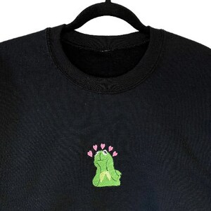 May include: Black crewneck sweatshirt featuring an embroidered Kermit the Frog design with pink hearts above his head. The frog is green with a light green belly. The sweatshirt is hanging on a black hanger.