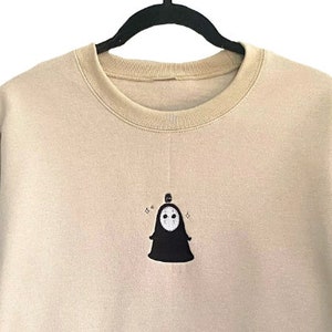 Op de afbeelding: Beige crewneck sweatshirt met een geborduurd zwart-wit karakterontwerp. Het ontwerp is gecentreerd op de voorkant. De sweatshirt hangt aan een zwarte hanger.