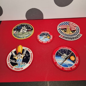 May include: Five embroidered patches featuring NASA space shuttle missions. The patches are circular and feature various space shuttle designs, astronaut names, and mission details. The patches are on a red background.