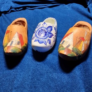 May include: Three wooden clogs, one with a blue and white floral design, and two with painted scenes of houses and windmills.