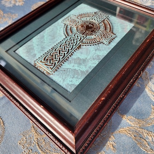 May include: A framed Celtic cross with intricate knotwork design in copper tones, set against a blue and white textured background. The frame is dark wood with a decorative edge.