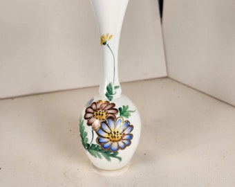 May include: A small white ceramic vase with a gold rim and floral designs. The vase features a blue, pink, and yellow flower with green leaves.