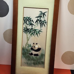 May include: A framed artwork featuring a panda bear eating bamboo. The panda is black and white, set against a backdrop of green bamboo and grass. The frame is a dark brown color, and the artwork is rectangular.