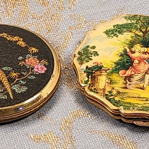 May include: Two vintage compact mirrors. One has a black background with a gold pheasant and floral design. The other has a gold background with a painted scene of a couple in a garden.