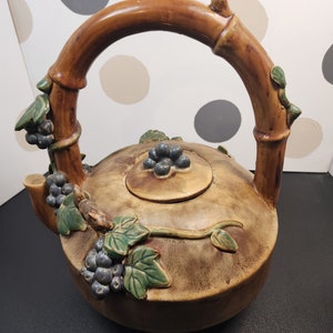 May include: A brown ceramic teapot with a bamboo-style handle and a vine design with green leaves and purple grapes. The teapot has a lid with a knob in the center.