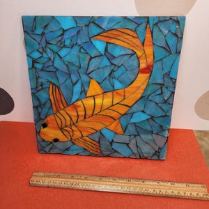 May include: A square mosaic artwork featuring an orange and yellow koi fish against a background of blue and turquoise glass pieces. The fish is detailed with black outlines. The artwork is approximately 12 inches wide.