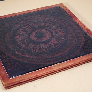 May include: Square wooden coaster with a dark, patterned center. The center design features a circular pattern in shades of brown and black, framed by a wooden border. The coaster is a decorative item for home use.