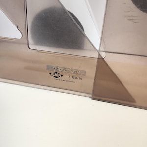 May include: A clear, geometric drafting tool made of acrylic. The tool has a brown tint and features the text "ALVIN T 160-14 MADE IN W-GERMANY". The design includes a circular shape and angular edges.