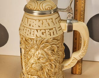 May include: Ivory-colored beer stein with a lion's head relief and the text "Afrika's Serengeti." The lid features a silver-colored metal rim and a decorative elephant figurine. The handle is cream-colored.