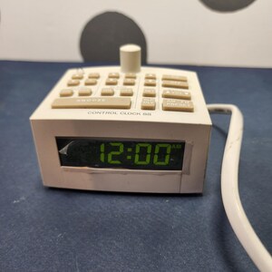 May include: A vintage white Control Clock 88 alarm clock with beige buttons and a digital display showing 12:00 AM. The clock has a cylindrical knob on top and a white cord.