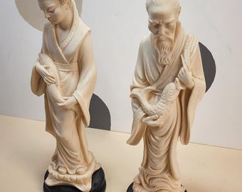 May include: Two ivory-colored statues of Asian figures, one male and one female, each holding a fish. The figures are on black circular bases. The statues have detailed facial features and traditional clothing. The background is a neutral color with a light-colored circle.
