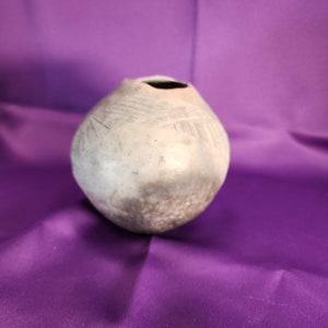 May include: A small, round, gray ceramic pot with a wide opening at the top. The pot has a textured surface and appears to be hand-made.