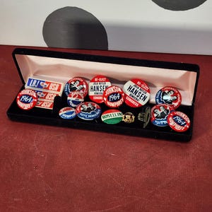 May include: A collection of vintage political campaign buttons and pins, including several with the text "Johnson 1964" and "Humphrey." The buttons are red, white, and blue, and are displayed in a black velvet box. Some buttons read "Re-Elect Hansen."
