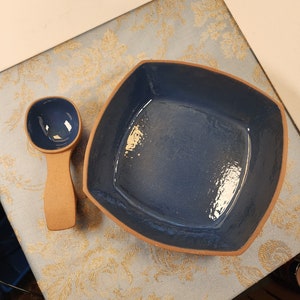 May include: A square blue ceramic serving dish with a wooden spoon. The dish has a slightly raised rim and a glossy finish.