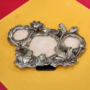 May include: Silver-toned metal picture frame with three oval openings. The frame is decorated with detailed frog and lily pad designs. The frame has a black stand and is set against a yellow and red background.