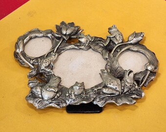 May include: Silver-toned metal picture frame with three oval openings. The frame is decorated with detailed frog and lily pad designs. The frame has a black stand and is set against a yellow and red background.