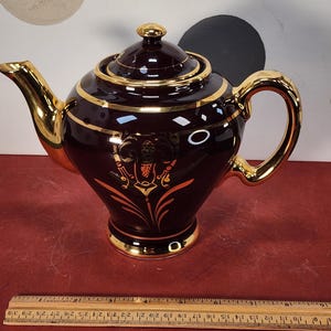 May include: A dark burgundy teapot with gold trim and accents. The teapot features a gold handle, spout, and lid. The design includes gold bands and a decorative emblem. The teapot is approximately 10 inches long.