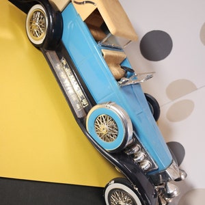 May include: A blue and black toy car with a tan top. The car has chrome accents and a detailed engine. The car is on a yellow and black surface.