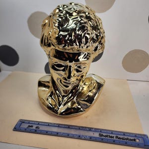 May include: A gold-colored bust of a classical figure, likely a sculpture, with detailed facial features and curly hair. The bust is made of a shiny material and is placed on a light brown surface. A blue ruler is placed in front of the bust.