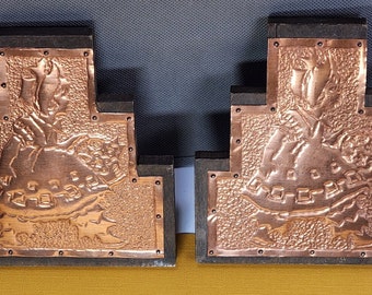 May include: A pair of decorative bookends made of wood and copper. Each bookend features a raised copper design of a stylized figure with a floral motif. The bookends are a dark brown color and have a rustic appearance.
