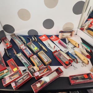 May include: A collection of vintage fishing lures in various colors and designs, some in original packaging. The lures include crankbaits, spoons, and other styles, with visible hooks and detailed paint jobs. The collection is displayed on a dark surface.