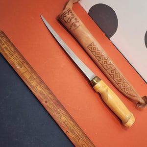 May include: A J. Marttiini Finland fillet knife with a silver blade, a light wood handle, and a brass bolster. It is accompanied by a tan leather sheath featuring embossed geometric patterns and the text J. Marttiini Finland.