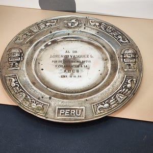 May include: A silver plate with intricate embossed designs and the word "PERU" at the bottom. The plate features text in Spanish, including the name "LORENZO VASQUEZ L." and the date "11-11-84". Decorative elements include stylized figures and patterns.