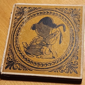 May include: A vintage book cover with a black and white illustration of a cartoon dog and a character with a black hat. The illustration is surrounded by a decorative gold frame.
