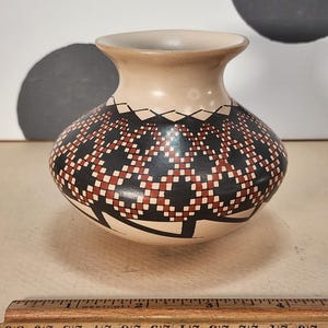 May include: A ceramic vase with a beige neck and a rounded body. The body features a geometric pattern in black, red, and white. The vase is approximately 4 inches tall.