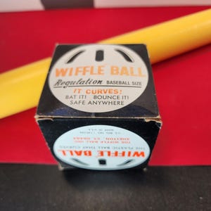 May include: A black box with a white Wiffle Ball logo and the text "Wiffle Ball Regulation Baseball Size" and "It Curves! Bat It! Bounce It! Safe Anywhere". The box is sitting on a red surface with a yellow object behind it.
