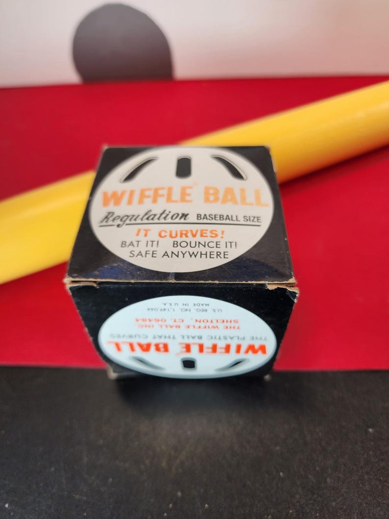 May include: A black box with a white Wiffle Ball logo and the text "Wiffle Ball Regulation Baseball Size" and "It Curves! Bat It! Bounce It! Safe Anywhere". The box is sitting on a red surface with a yellow object behind it.