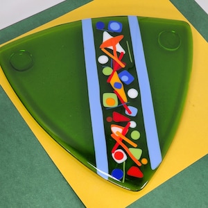 May include: A green glass triangular plate with a blue and white stripe down the center. The stripe is decorated with colorful geometric shapes, including red, orange, yellow, green, blue, and white.