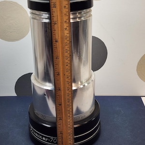 May include: A silver Prominar-70 telescope with a black base and top. The telescope is cylindrical and measures approximately 10 inches tall. The text "Prominar-70" and "104 mm" are visible on the base.